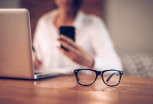 close up of eye glasses on a desk with woman on phone in background