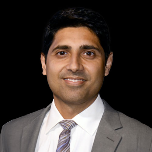Professional headshot of ophthalmologist Dr. Amit Sangave, in a tan blazer and striped tie, smiling in front of a dark backdrop.