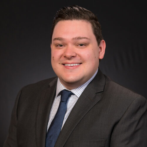 Professional headshot of optometrist Dr. Cody Frank, wearing a dark suit and patterned tie, smiling brightly against a gray background.