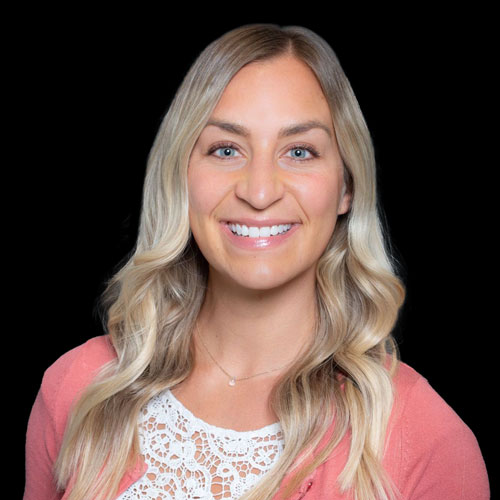 Professional headshot of optometrist Dr. Jessie LaLonde, wearing a coral cardigan and floral blouse, smiling against a black background.