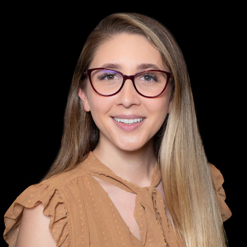 Professional headshot of optometrist Dr. Patrizia Colmenares, with long blonde hair and glasses, wearing a tan blouse against a black background.