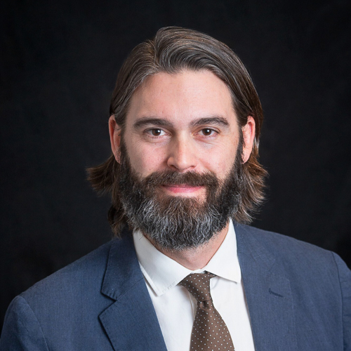 Professional headshot of ophthalmologist Dr. Brendan Girschek, with long hair and beard, wearing a brown suit and tie, posed against a dark background.