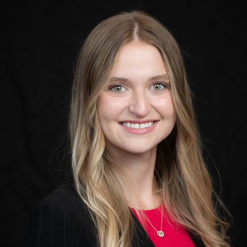 Professional headshot of optometrist Dr. Emily Brill, with long blonde hair and a pink blouse, smiling against a black background.