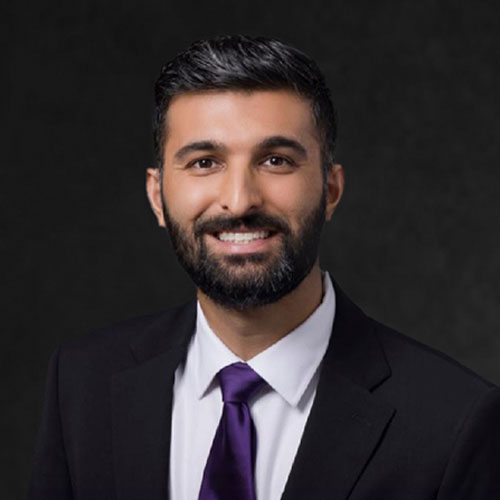 Dr. Kanwardeep Sidhu, optometrist, smiling warmly while wearing a dark suit and a purple tie, against a black neutral background.