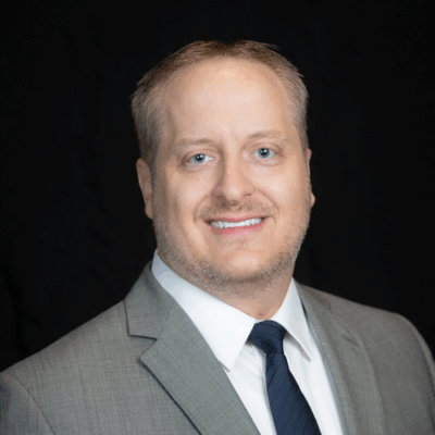 Professional headshot of Dr. Shane Theobald, optometrist at Southwestern Eye Center, wearing a light grey suit and dark blue tie, smiling confidently against a dark background.