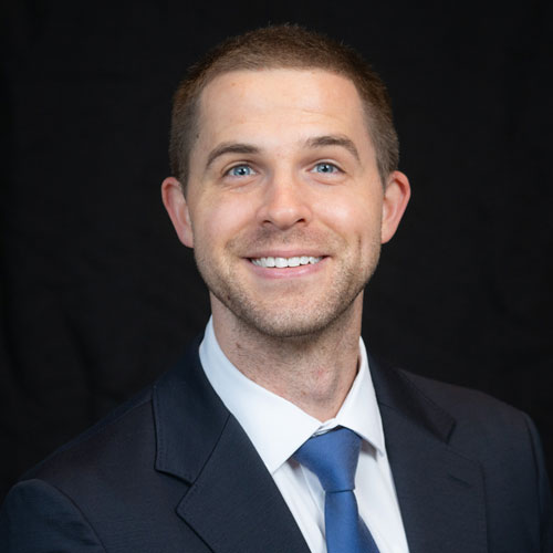 Professional headshot of optometrist Dr. Alex Christensen, wearing a navy suit and light blue shirt, smiling warmly in front of a dark backdrop.