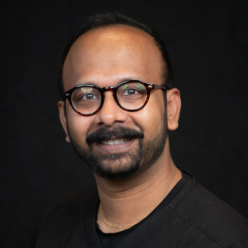 Professional headshot of ophthalmologist Dr. Tamzid Rahman, wearing round glasses and a black shirt, smiling in front of a black backdrop.