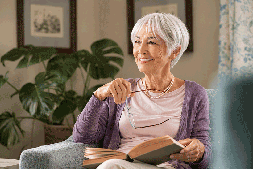 Senior woman holding glasses while reading at home, representing common causes of dry eyes at Southwestern Eye Center.