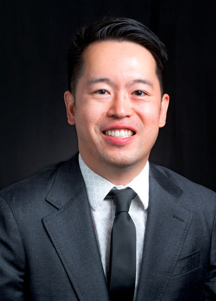 Portrait of a smiling man in a dark suit and tie against a dark background.