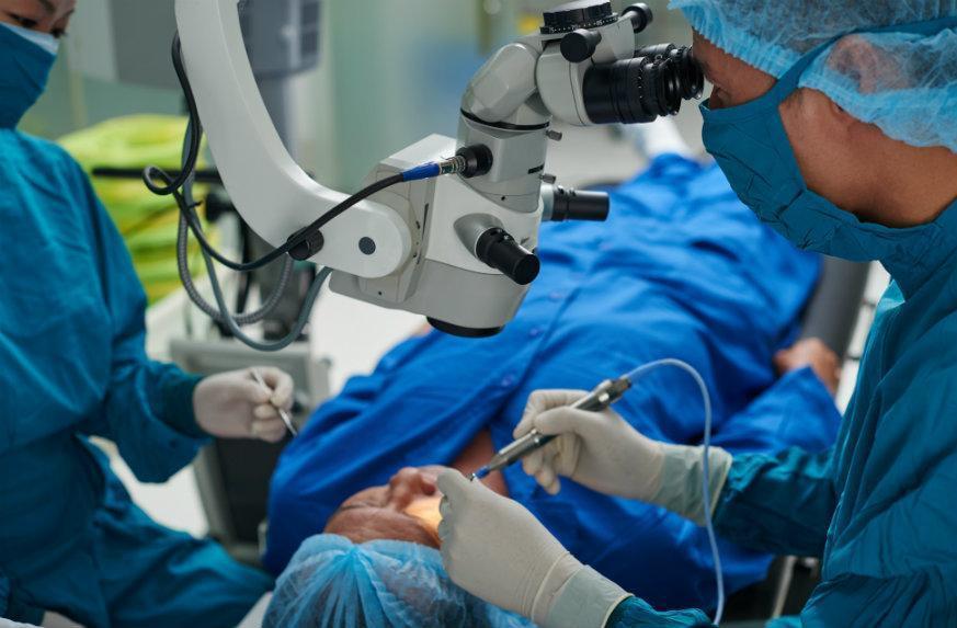 Surgeons in blue scrubs operate under a surgical microscope in an operating room.