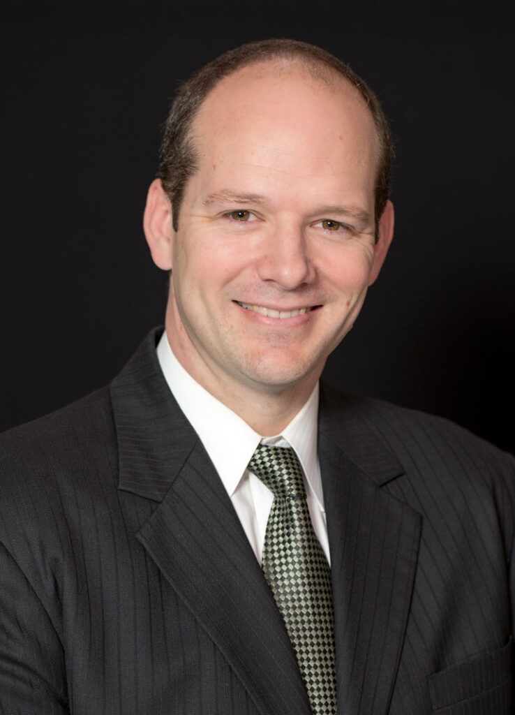 Professional headshot of optometrist Dr. Jordan Bluth, in a dark suit and patterned tie, smiling against a black background.