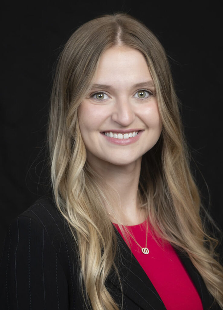 Professional headshot of optometrist Dr. Emily Brill, with long blonde hair and a red blouse, smiling against a black background.