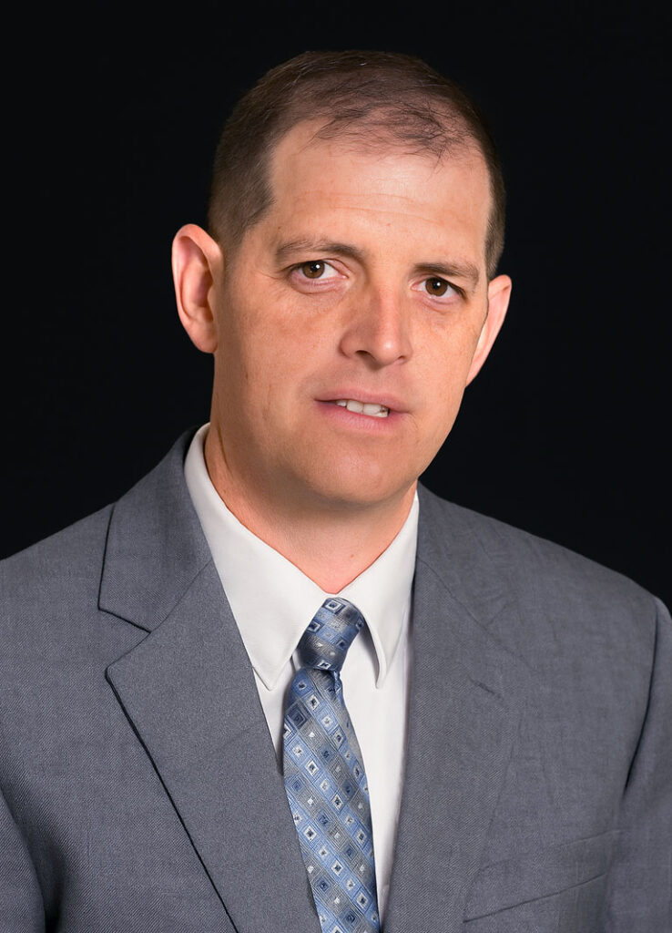 Professional headshot of optometrist Dr. James Hooper, in a light gray suit and patterned tie, posed against a black background.