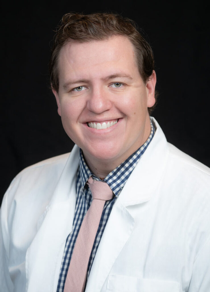 Smiling man in a white lab coat, checkered shirt, and pink tie against a dark background.