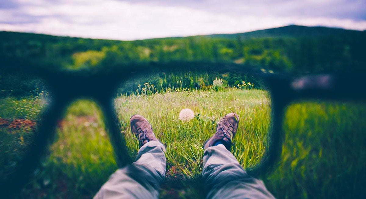 View of a person lying in a grassy field seen through the blurred frame of eyeglasses, with the scene in front more focused in the center and softer at the edges, illustrating how peripheral vision can look and supporting an educational blog from Southwestern Eye Center