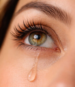 Close-up of a hazel and green eye with long eyelashes looking upward as a single tear rolls down the cheek, illustrating why do we cry and highlighting tear function and eye health care at Southwestern Eye Center