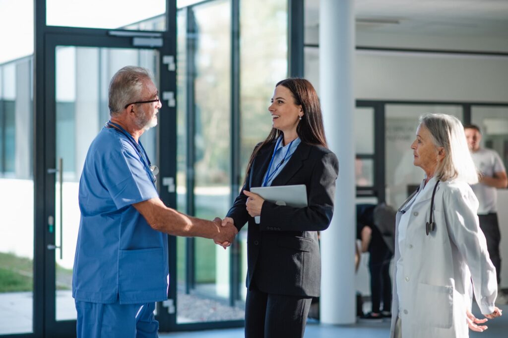 A doctor in blue scrubs shakes hands with a businesswoman in a dark suit as a colleague in a light blazer looks on in a modern lobby.