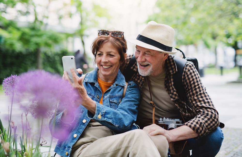 An elderly couple in a sunny park take a selfie with a smartphone, with purple flowers in the foreground.