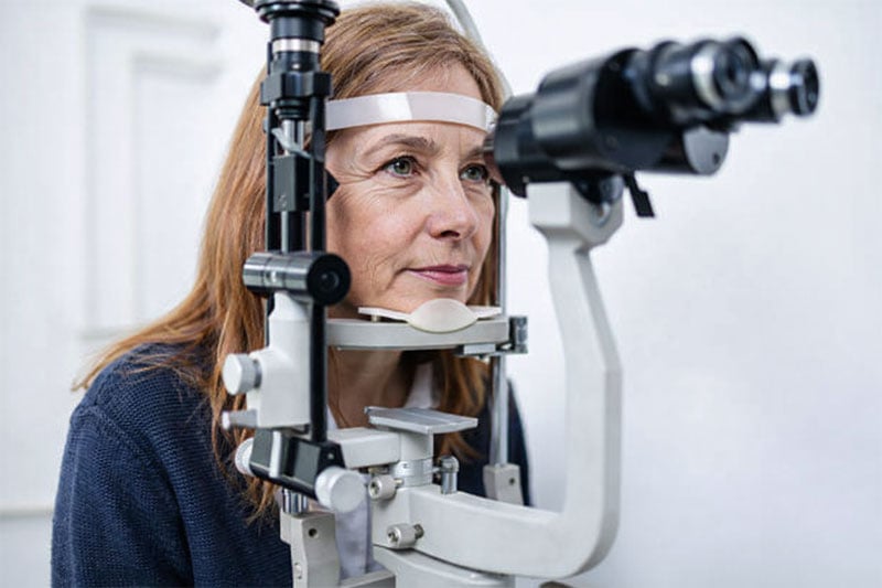 Older woman in her 60s undergoing an eye exam using a slit lamp machine, resting her chin on the support while looking into the microscope in a clean, modern eye care clinic.